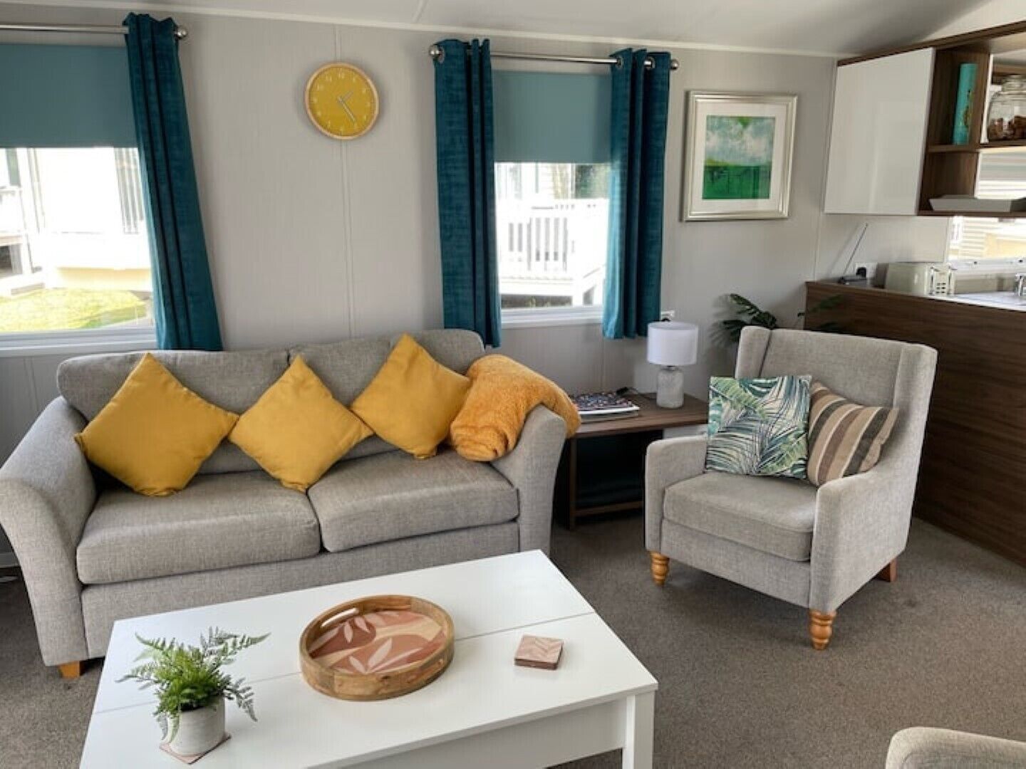 A living room with a grey sofa, a white coffee table, and a grey armchair. There is a clock on the wall and a framed picture above the sofa.