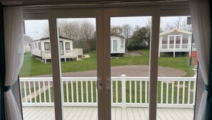 A view through a sliding glass door of a deck area with a white railing, leading to a grassy yard with two mobile homes. - 6 Berth Comfy Homely Caravan, Dog Friendly (Burgh Castle)