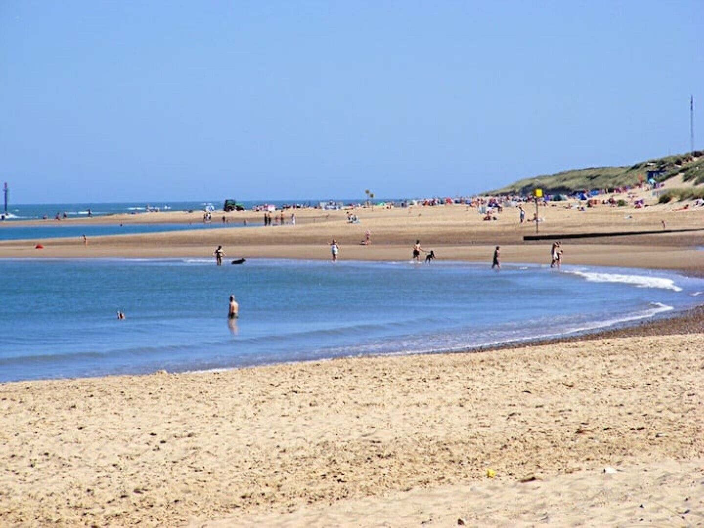 Une vaste plage de sable fin bordée d’une eau d’un bleu limpide, où l’on voit des gens se promener, et une colline lointaine parsemée de constructions.