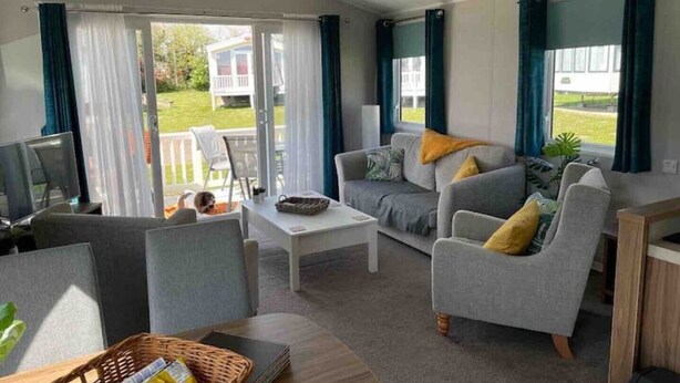 A spacious living room with a sofa, armchairs, a coffee table, and a TV. There is a sliding glass door leading to a balcony with outdoor furniture. - 6 Berth Comfy Homely Caravan, Dog Friendly (Burgh Castle)