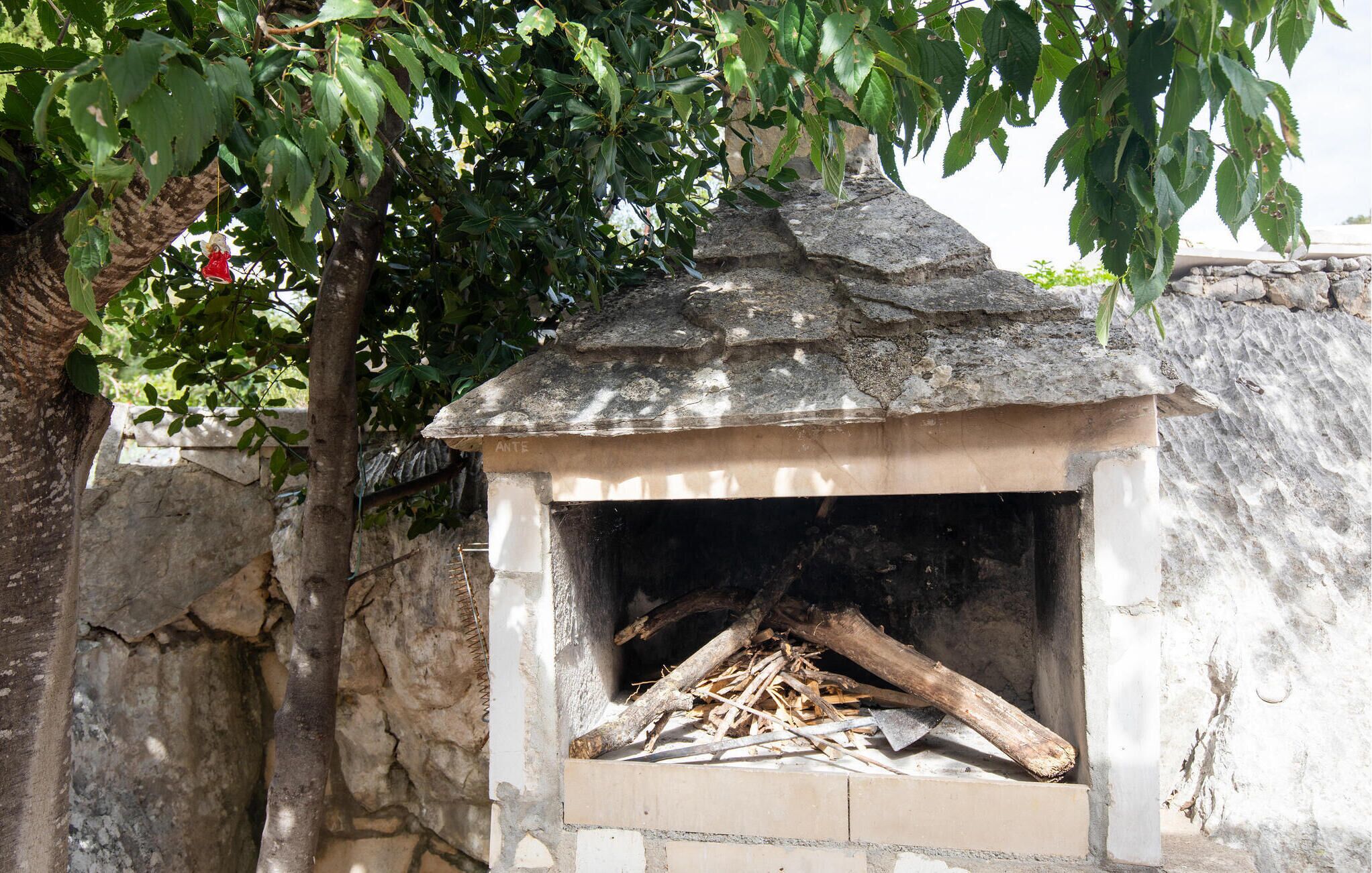 Una xemeneia exterior de pedra, amb teulada de rajola, envoltada per una paret de pedra i un arbre amb fulles verdes.