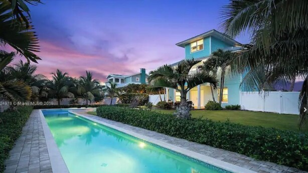 A swimming pool with a paved walkway, surrounded by well-maintained greenery and palm trees, leading to a building with a light blue exterior. - Stunning 4/3 w/ Heated Pool, Water View & Arcade! (Jensen Beach)