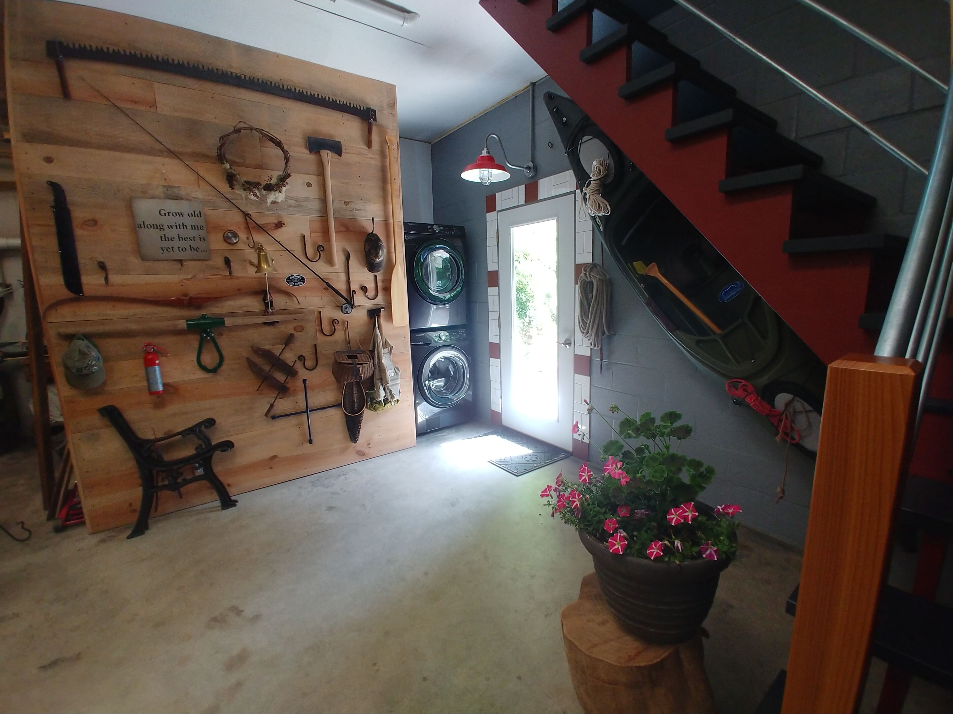 Une pièce avec un mur en bois où sont accrochés divers outils et des crochets, un lave-linge, un escalier et une plante en pot ornée de fleurs.