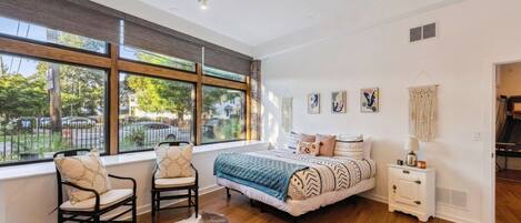 A bedroom with a large window, a bed with patterned bedding, a small bedside table, and a chair.