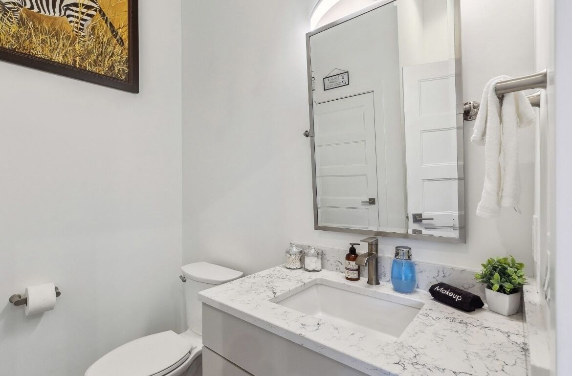 A bathroom with a white toilet, a sink with a marble countertop, a mirror above the sink, and a framed picture on the wall.