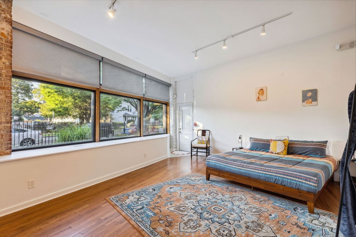 A bedroom with a large window, a bed with patterned bedding, a rug, and a chair.