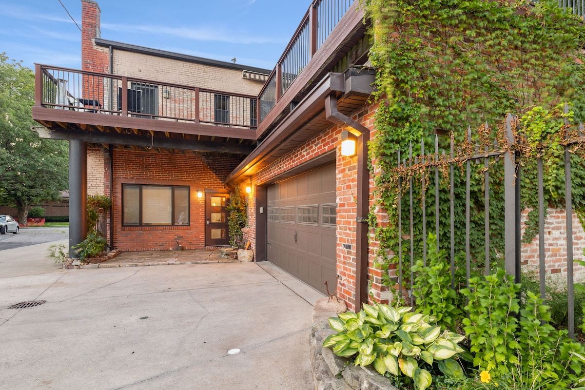 A two-story residential building with a garage, a balcony, and a garden with plants.