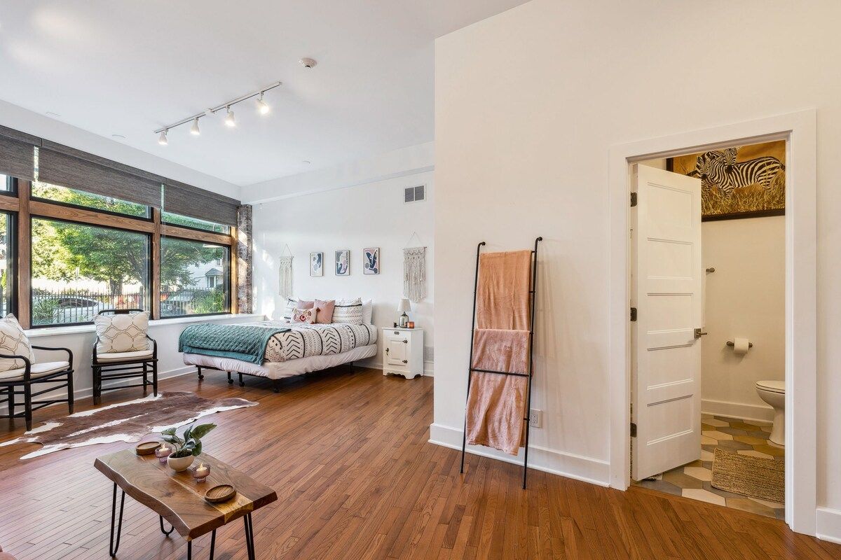 Une chambre spacieuse avec une grande fenêtre, un lit recouvert d’une couvre-lit à motifs, une table de chevet et une petite table de nuit.