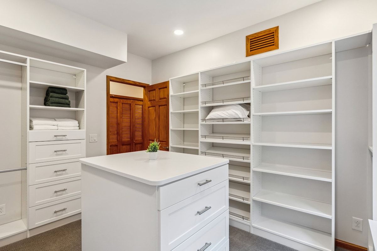 Ein begehbarer Kleiderschrank mit offenen Regalen, einer zentralen weißen Kommode und einer Holztür.