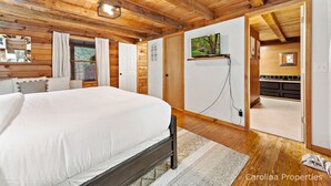 A bedroom with a large bed, wooden ceiling beams, a TV mounted on the wall, and a view into the kitchen. - Pet Friendly, Resort Access, Private! - Catahoula Cabin by Carolina Properties (Lake Lure)