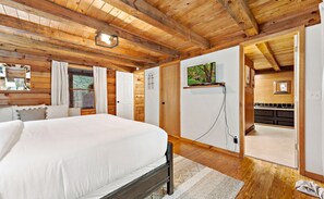 Un dormitorio con una cama grande, vigas de madera en el techo, un televisor montado en la pared y vistas a la cocina.