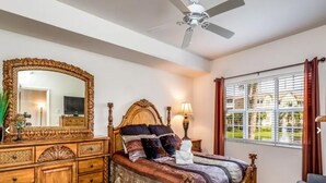 Un dormitorio con un espejo grande y decorativo, una cama de madera con muchos cojines, un ventilador de techo y una ventana con persianas.