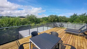 A wooden deck with a glass balustrade, outdoor seating, and a view of greenery.