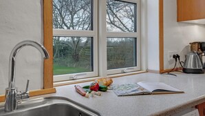 A kitchen with a stainless steel sink, a window with a view of trees, and a countertop with a loaf of bread, vegetables, and a cookbook.