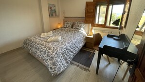 A bedroom with a bed, bedside table, lamp, chair, desk, and window.