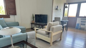 A modern living room with a sofa, armchair, and a glass-top coffee table. A television is on a stand, and there is a potted plant on a side table.