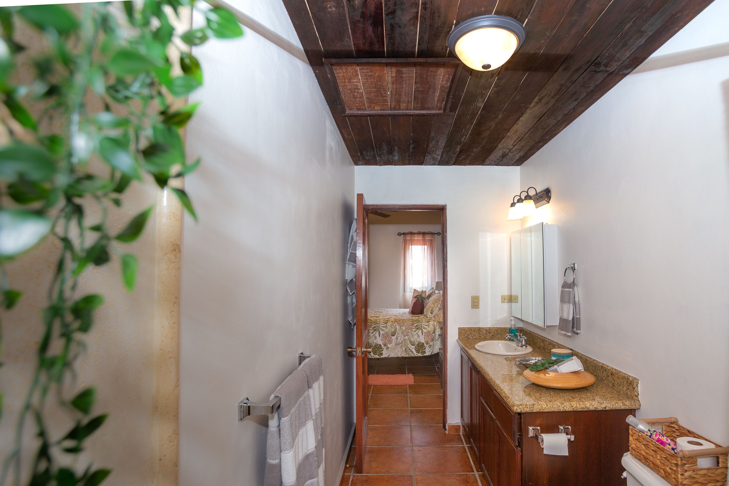 A narrow hallway leading to a bedroom with a bed, a bathroom with a sink and mirror, and a ceiling fan.