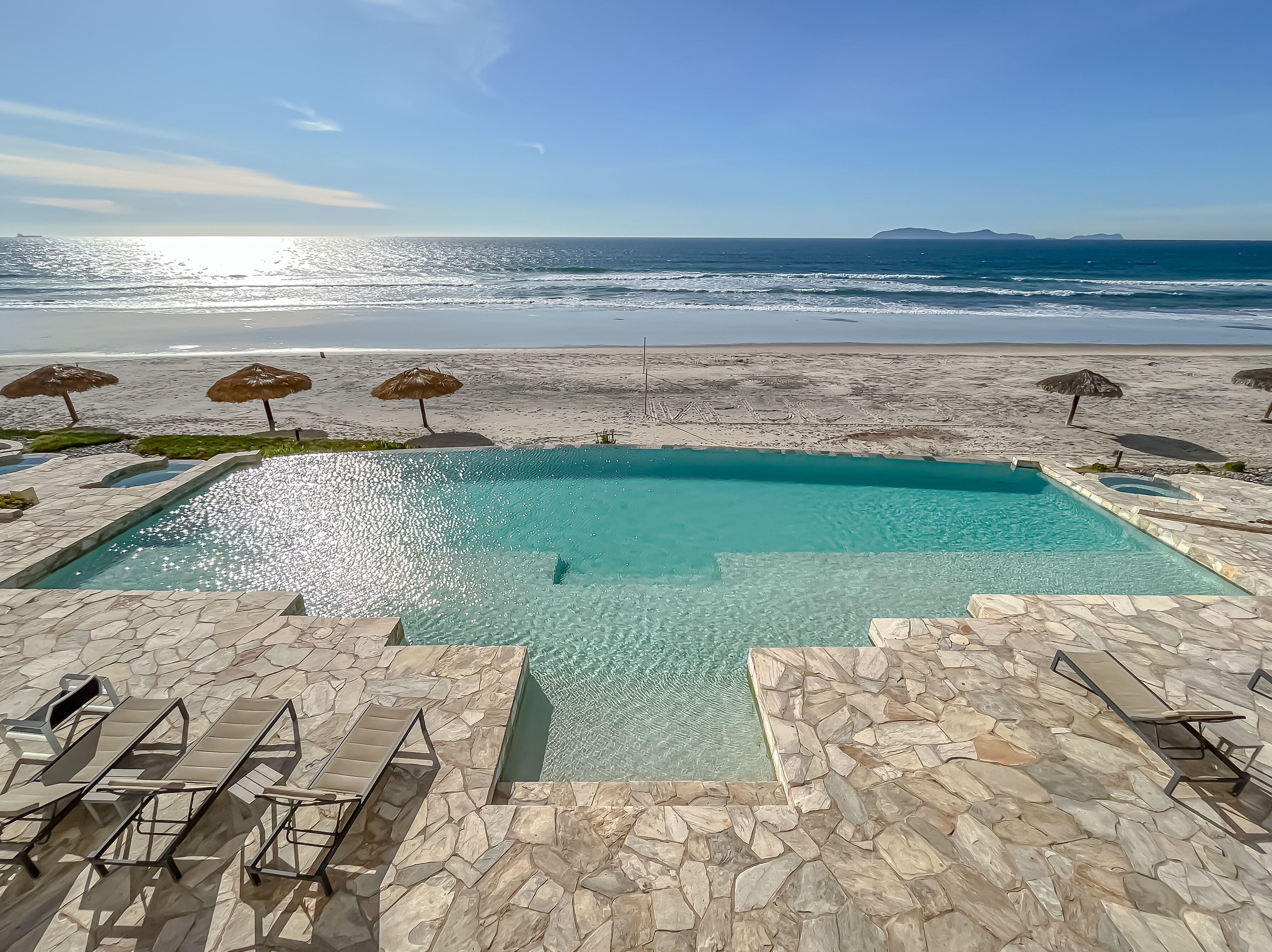 Piscina con vista al océano, rodeada de baldosas de piedra y sillones de descanso.