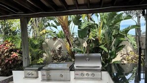 An outdoor kitchen with a gas stove, grill, and storage cabinets, surrounded by lush greenery. - The WaterSun Oasis - Luxury Private Pool & Spa (Naples)
