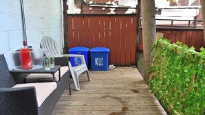 Een houten terras met buitenmeubilair, twee blauwe vuilnisbakken en een houten omheining.