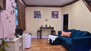 Een woonkamer met een blauwe bank, een klein tafeltje en een televisie. Twee schilderijen hangen aan de muur en links staat een plant.
