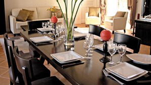 A dining area with a long table set for a meal, surrounded by chairs. There are vases with flowers and a fruit bowl on the table.
