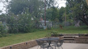 Un patio al aire libre con una mesa y sillas de metal, rodeado por un muro de piedra y follaje.