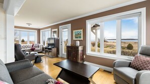 A spacious living room with a dark wooden coffee table, a grey sofa, and a grey armchair. There is a television on a wooden stand, and a cabinet with decorative items. Large windows offer a view of a landscape with trees and a body of water. - Spacious 4 bedrooms Villa, 30 min from Moncton (Cocagne)