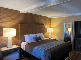 Quarto de hotel com cabeceira em padrão de tijolos, duas luminárias de cabeceira, cama com roupa de cama branca e azul, e mesa de cabeceira com gaveta.