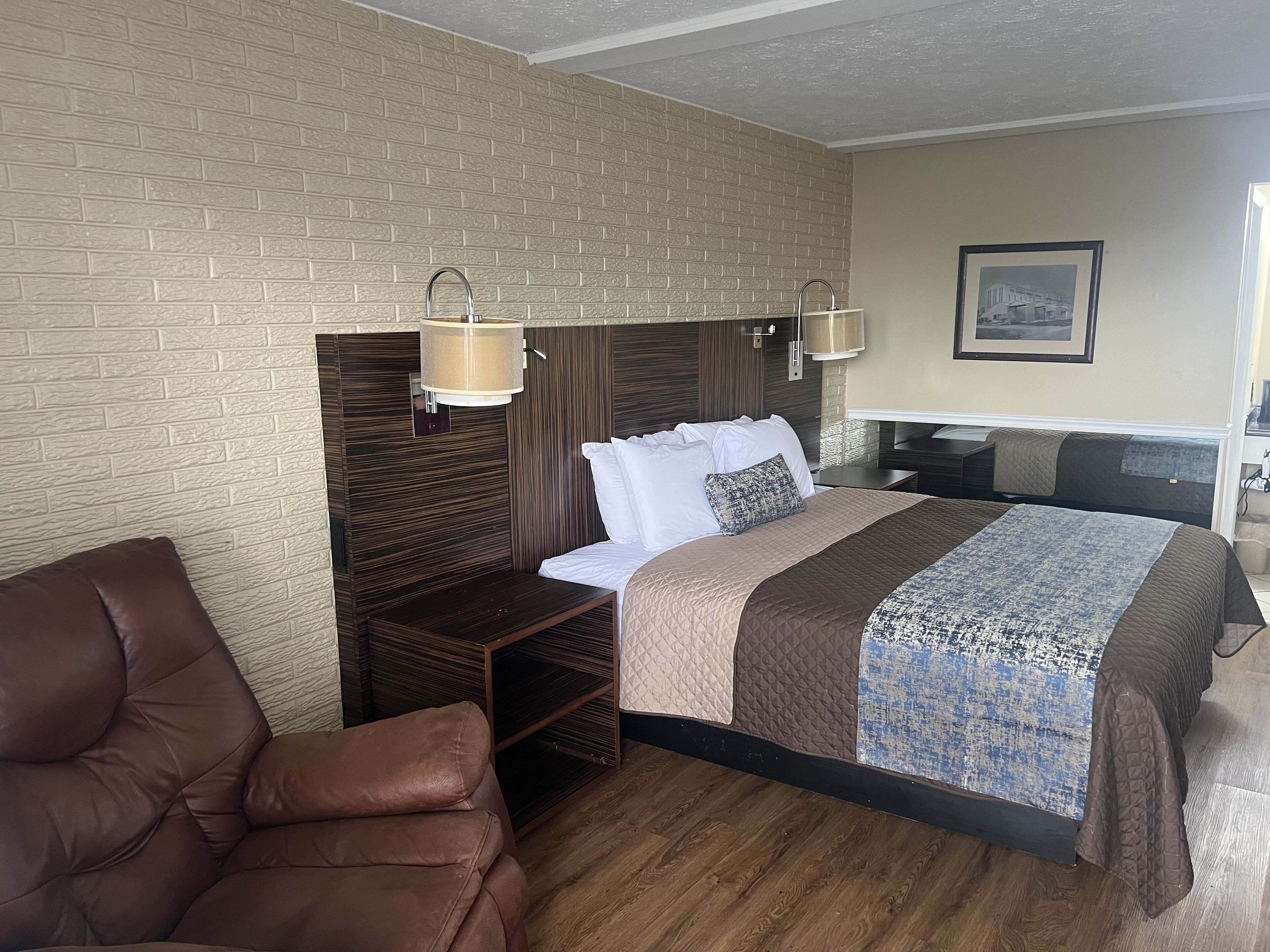 A hotel room with a brick-patterned wall, a bed with a patterned bedspread, a brown leather chair, a wooden nightstand, and a framed picture on the wall.