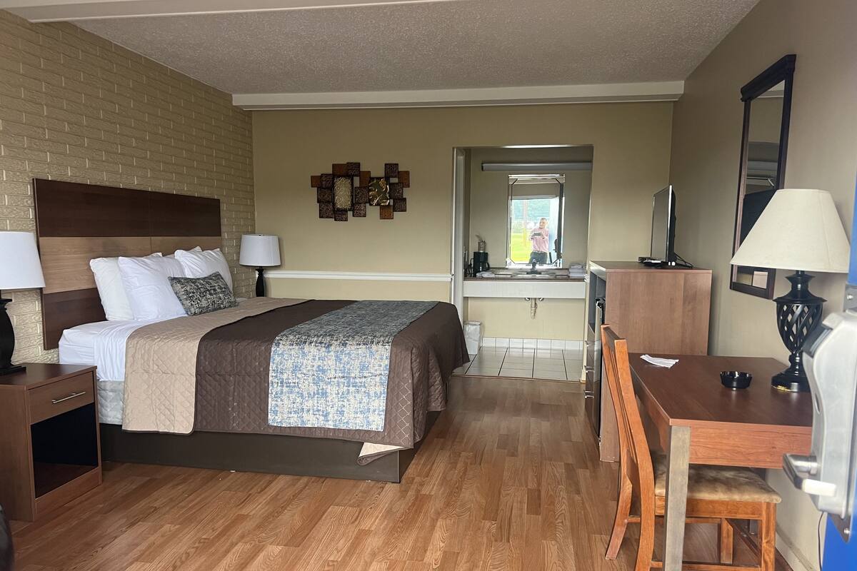 Quarto de hotel com cama, mesas de cabeceira, escrivaninha, cadeira, espelho e banheiro visível por uma porta aberta.