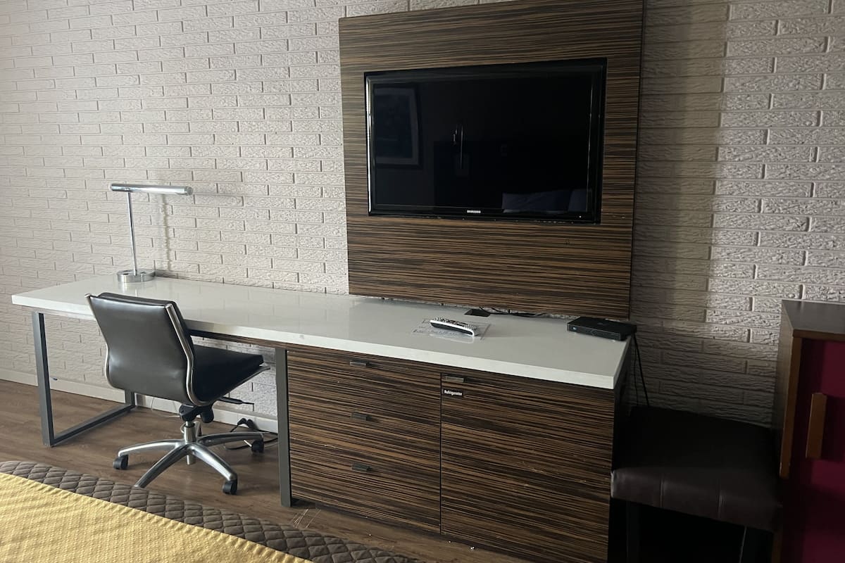 Escritório moderno com televisão de tela plana fixada em parede com textura de tijolos, uma mesa branca com cadeira e uma gaveta refrigerada embutida.