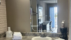 A bathroom with a marble countertop, a sink, a mirror, a towel dispenser, and a coffee maker. - Towne Inn (Sylacauga)