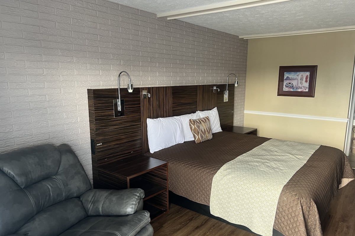 Quarto de hotel moderno com parede em padrão de tijolos, uma cama grande com colcha marrom, um sofá de couro cinzento e piso de madeira.