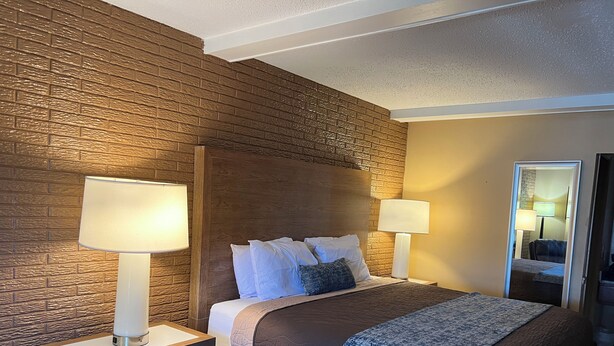 A hotel room with a brick-patterned wall, a wooden headboard, a bed with white and blue bedding, two bedside lamps, and a nightstand with a drawer. - Towne Inn (Sylacauga)