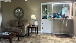 A living room with a sofa, coffee table, lamp, and a door leading to a kitchen area. - Towne Inn (Sylacauga)