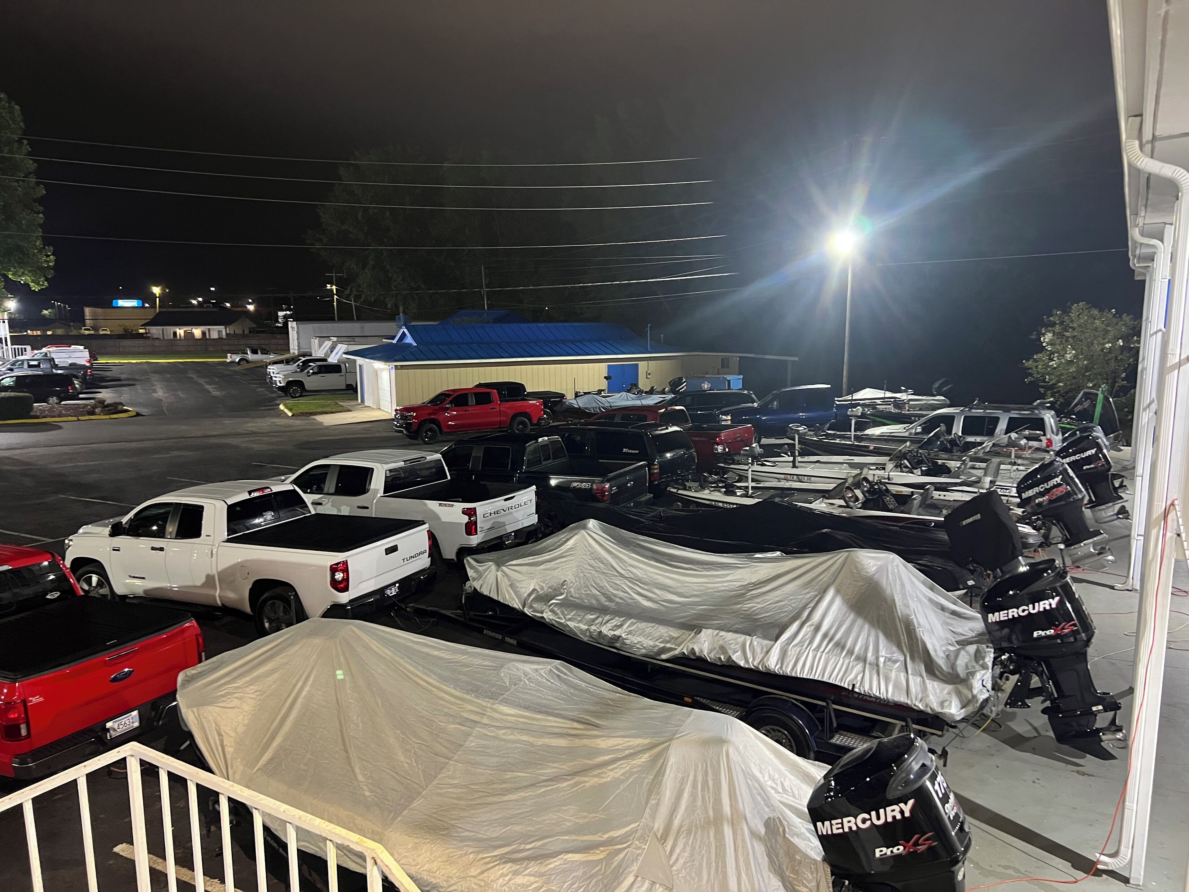 A parking lot with various vehicles, including trucks and covered boats, under bright lights at night.