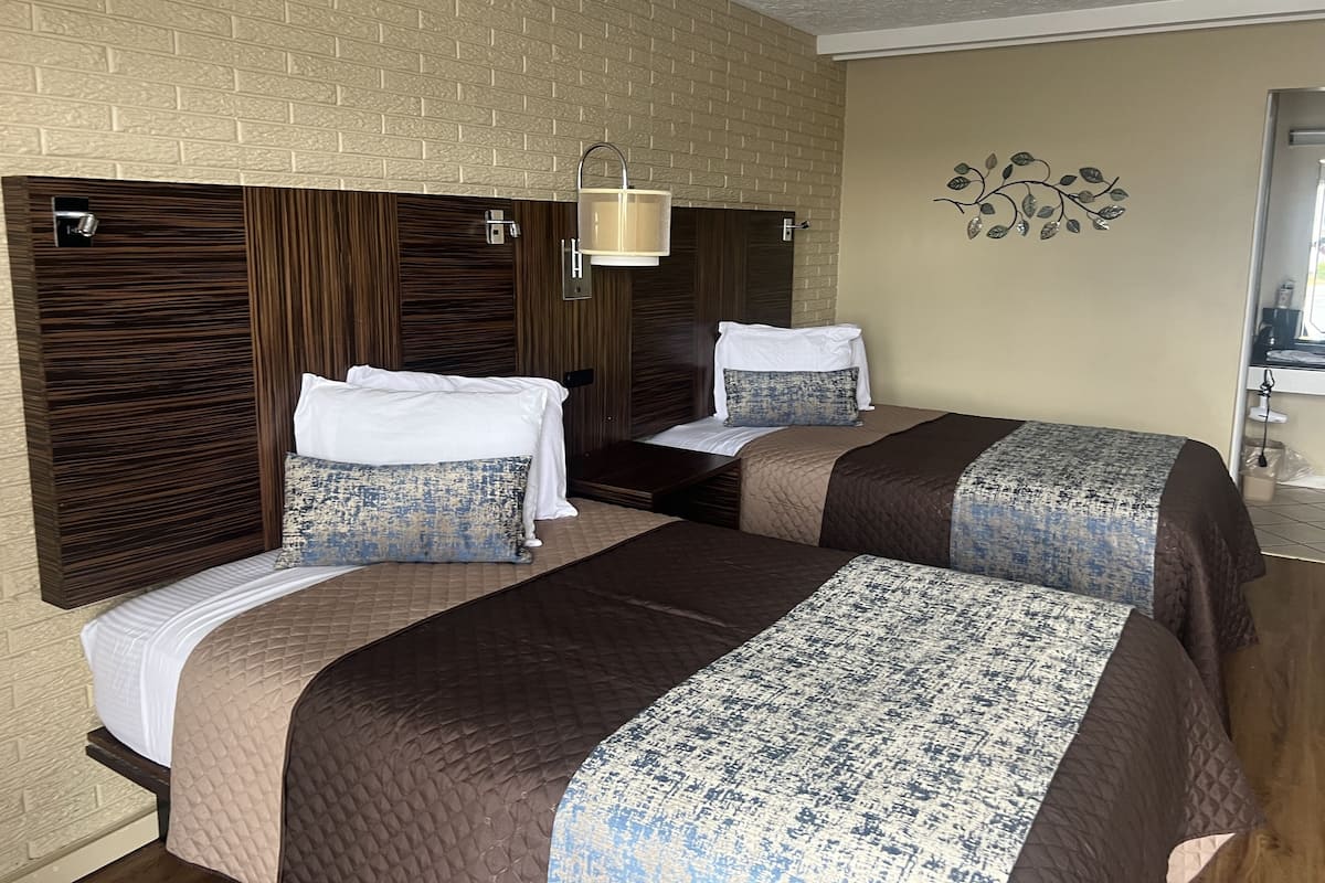 Quarto de hotel com duas camas, uma parede de tijolos, cabeceira de madeira, luminária fixada na parede e um desenho decorativo de folha.