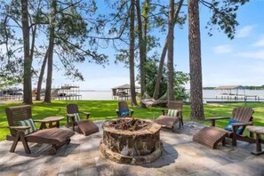 An outdoor seating area with wooden chairs and a central fire pit, surrounded by trees and a view of a body of water.