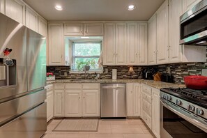 A kitchen with white cabinets, a stainless steel refrigerator, a dishwasher, a stove, and a window with a view of greenery.