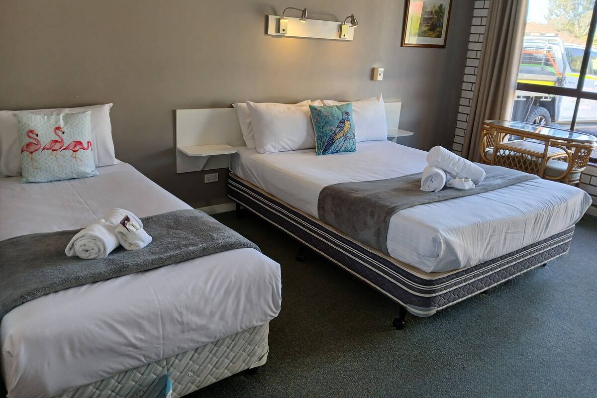 Habitación de hotel con dos camas, cada una con ropa blanca de cama y almohadas, una manta gris y una toalla enrollada. Hay una pequeña mesa con una silla, una lámpara de pared y un cuadro enmarcado.