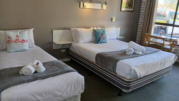 Habitación de hotel con dos camas, cada una con ropa blanca de cama y almohadas, una manta gris y una toalla enrollada. Hay una pequeña mesa con una silla, una lámpara de pared y un cuadro enmarcado.