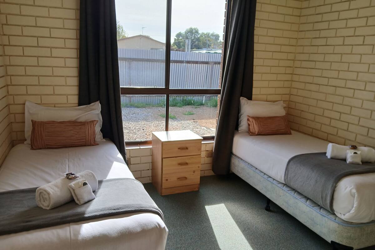 Una habitación con dos camas individuales, un pequeño aparador de madera y una ventana que da a un edificio y una valla.