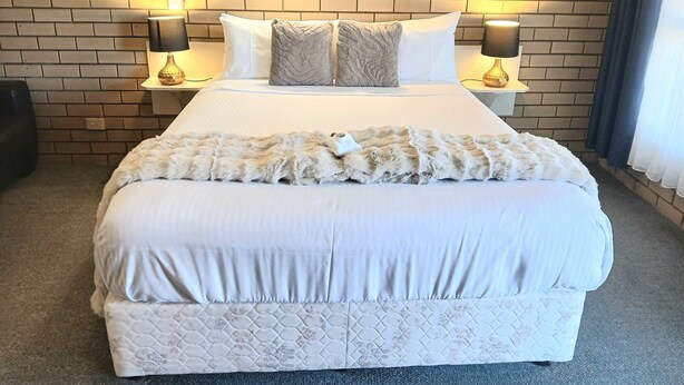 A hotel room with a brick-patterned wall, a large bed with white bedding, two bedside lamps, and a fluffy throw blanket. - Birch Motel Tocumwal (Tocumwal)