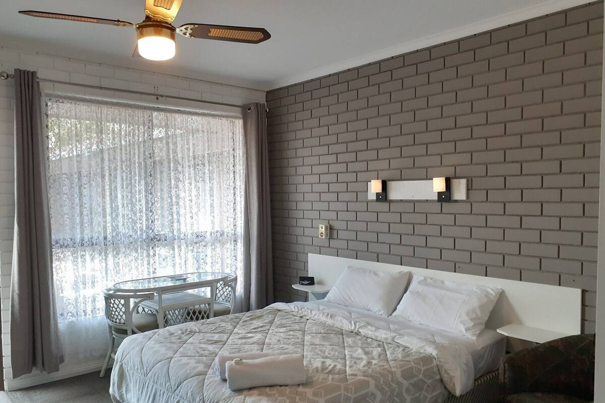 Habitación de hotel con cama, ventilador de techo, ventana con cortinas, una mesita y pared con diseño de ladrillos.