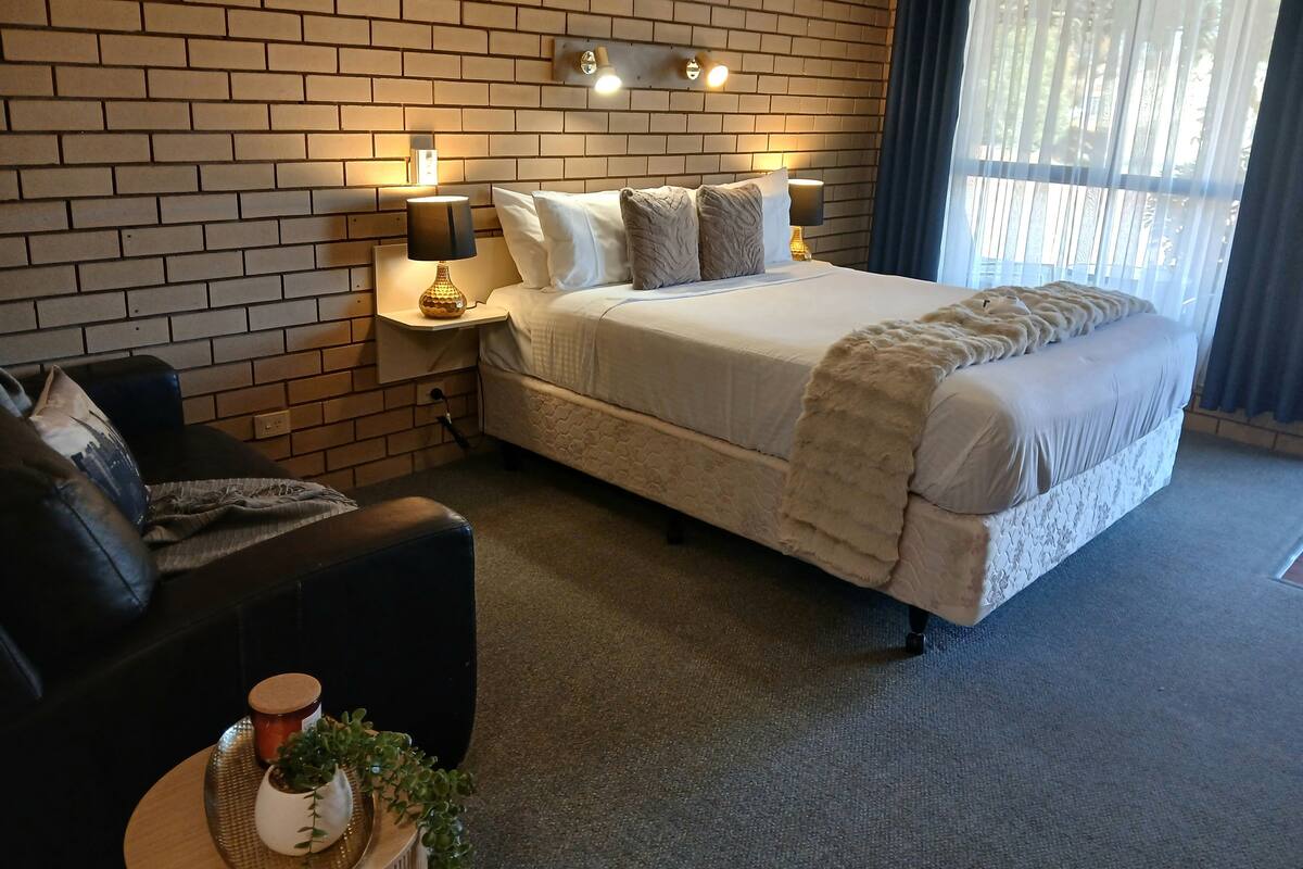Un dormitorio ordenado con pared de ladrillo, una cama grande con manta suave, una mesita de noche con lámpara y una pequeña mesa auxiliar con una planta.
