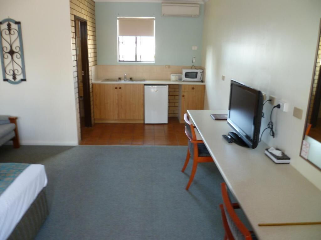 A compact room with a bed, a desk with a computer, a microwave, and a small kitchenette with a sink and a refrigerator.