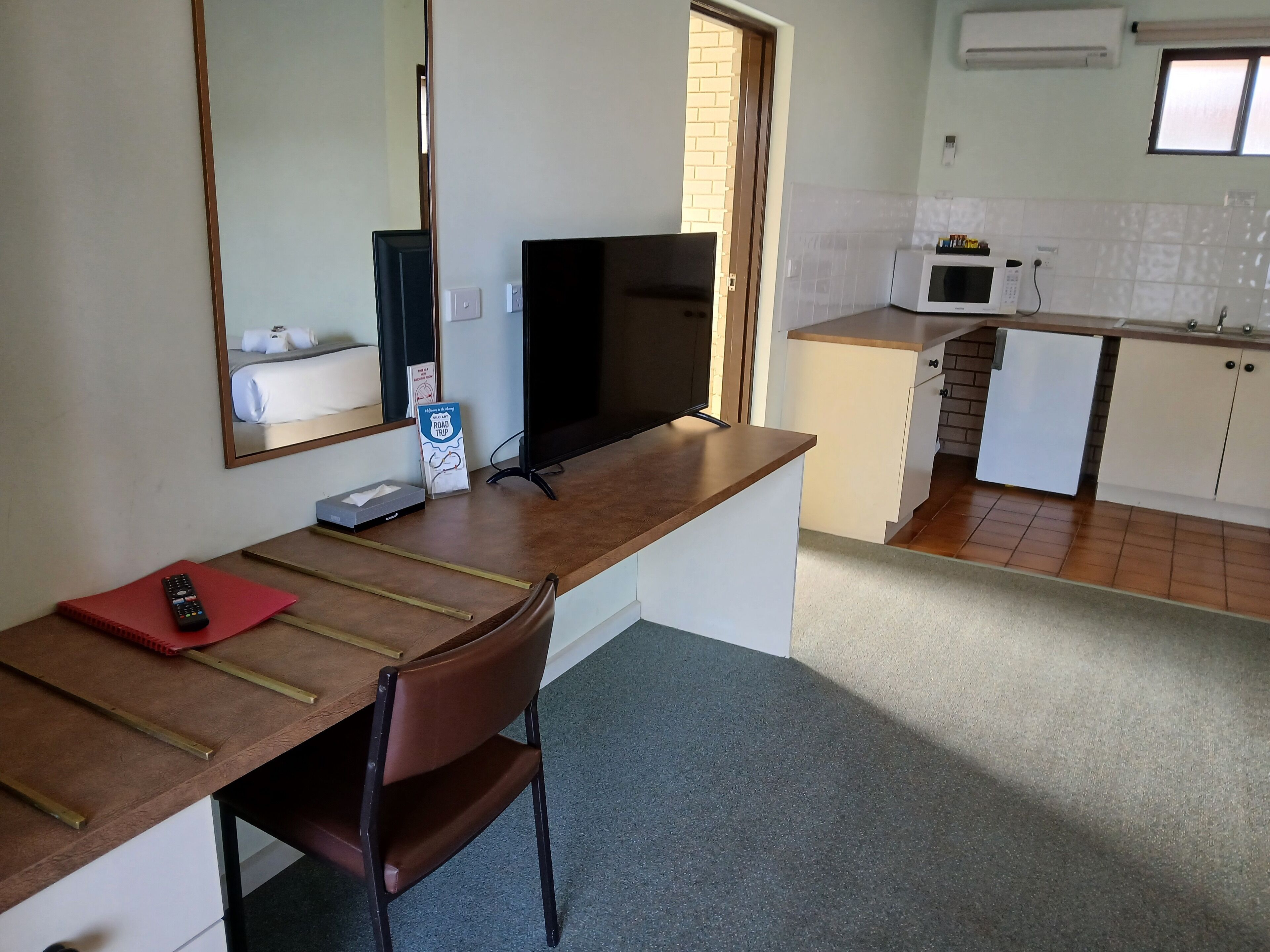 A compact hotel room with a kitchenette, a wooden desk with a chair, a television, and a mirror.