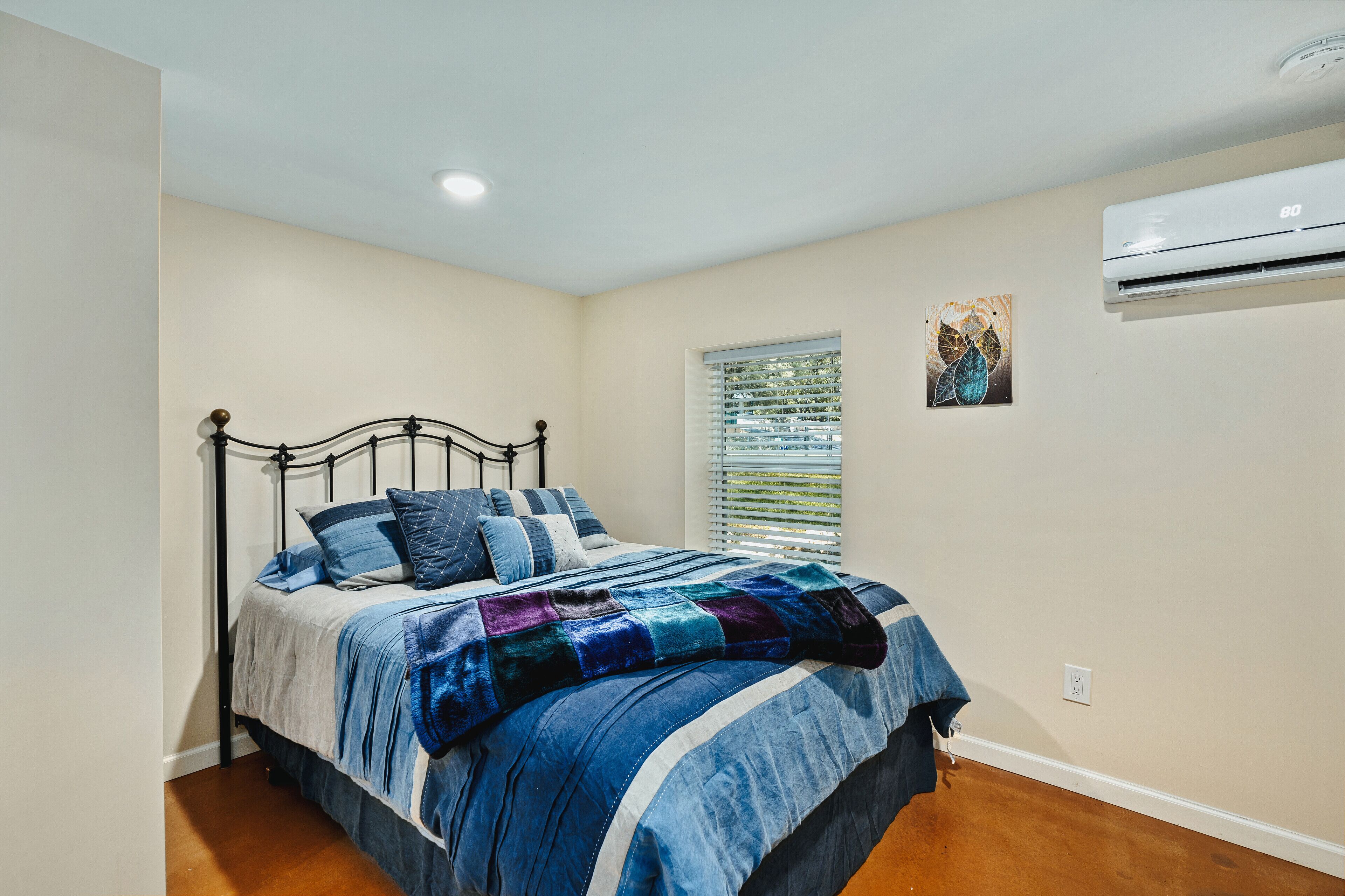 A bedroom with a bed, a window with blinds, an air conditioning unit, and a small painting on the wall.