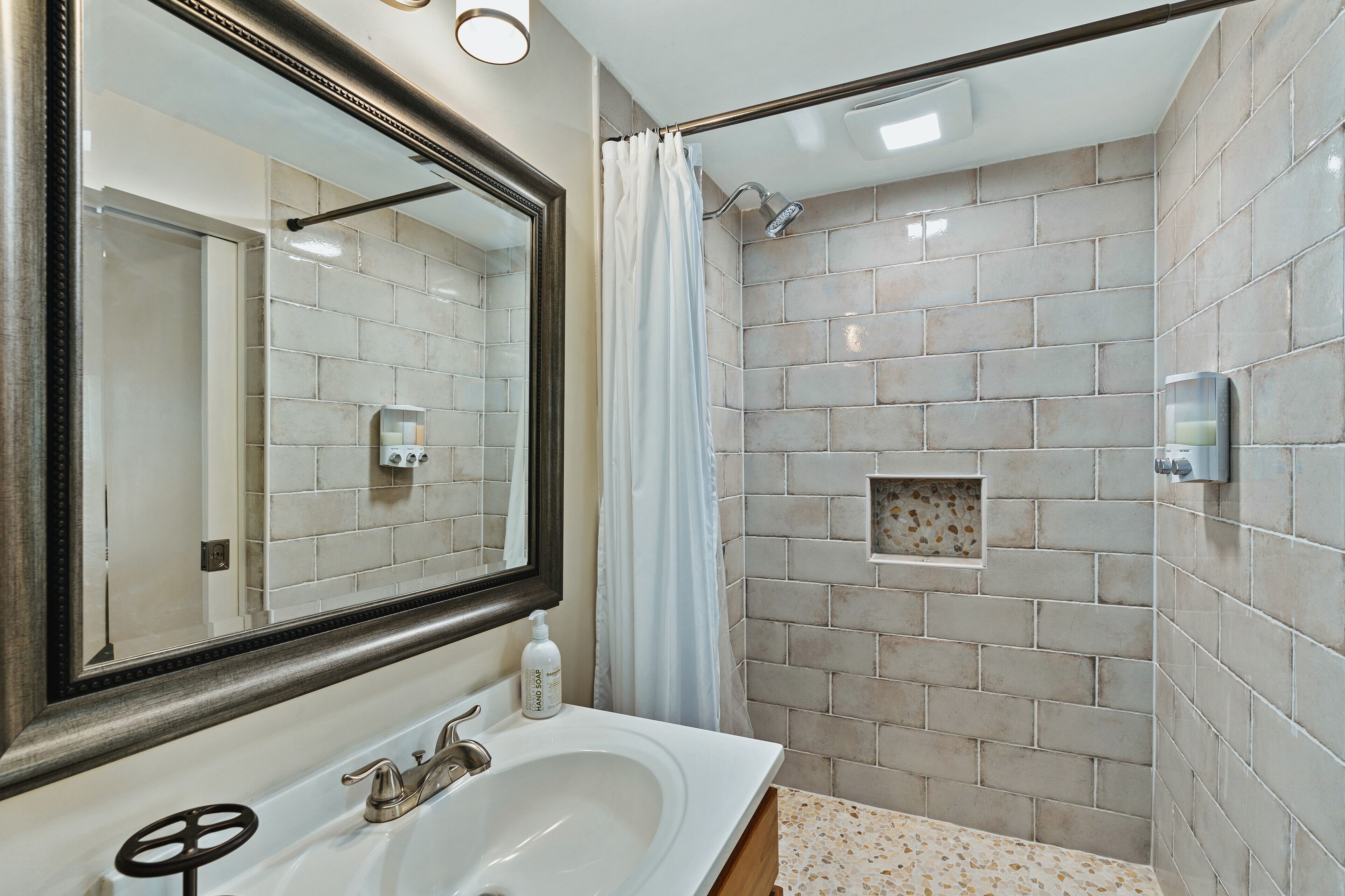 Un baño con un lavabo blanco, un espejo grande y una ducha con azulejos color beige.